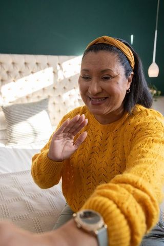 Mature woman waving during virtual call sitting on bed in mustard-yellow knit sweater