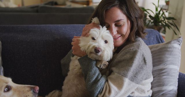 Woman sitting on comfortable couch hugging her white fluffy dog with another dog looking in background. Could be used to illustrate themes of pet love, family bonding, relaxation, or living room decor.