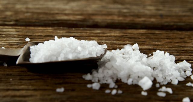 Coarse sea salt spills from a small metal scoop onto a rustic wooden table. The coarse, crystalline texture of the salt contrasts with the grainy wood surface, highlighting the natural and earthy feel. This image is ideal for use in culinary blogs, recipe websites, or food-related articles to emphasize organic ingredients and traditional cooking methods. It can also be used in advertisements for gourmet salts or kitchen supplies.