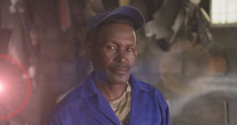 Veteran mechanic standing in blue coveralls in dim workshop with window light flare