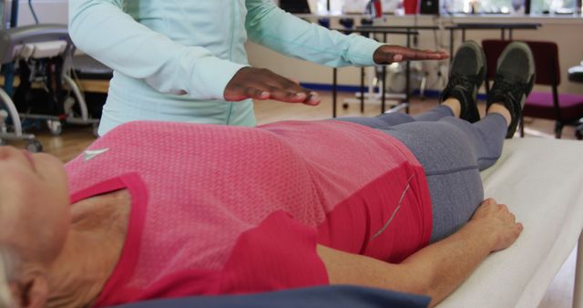 This image shows a healer performing a reiki therapy session on an elderly woman who is lying on a table. The elderly woman is wearing a pink top and blue pants. The healer's hands hover above her body, indicating energy healing. This image can be used for promoting alternative medicine practices, holistic healing, wellness retreats, and spirituality or relaxation techniques.