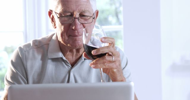 An elderly man sits at home, enjoying a glass of red wine while using his laptop. This image can be used to represent senior lifestyle, hobbies, retirement activities, relaxation, and online engagement among older adults. It is suitable for articles or promotions about senior living, health and well-being for elderly, technology use among seniors, or leisure activities.