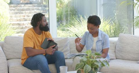 African couple planning finances at home using tablet and writing in notebook on sofa