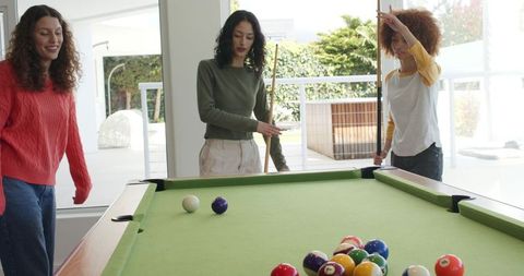 Diverse women playing pool in bright modern lounge with green felt table and cues