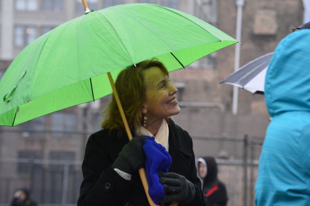A woman is smiling while holding a green umbrella on a rainy day in a city. This image is suitable for use in weather-related articles, urban lifestyle blogs, or marketing materials related to umbrellas and rain gear. It conveys a positive mood despite the rainy weather.
