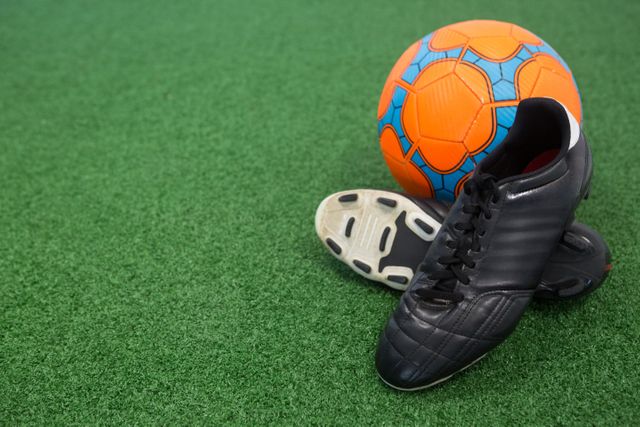 Close-up view of a football and cleats on artificial grass. Ideal for use in sports-related content, advertisements for athletic gear, soccer training materials, and articles about football equipment.
