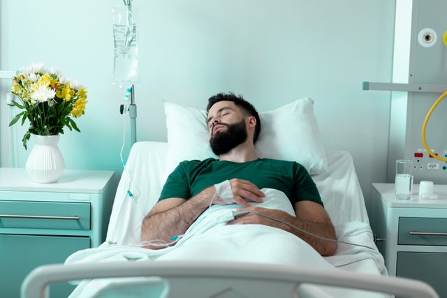 Bearded male patient lying asleep in a hospital bed with an IV drip attached to his hand. A vase of flowers is on the bedside table, adding a touch of comfort to the clinical environment. This image can be used for healthcare and medical service promotions, patient care brochures, hospital websites, and articles about recovery and medical treatments.