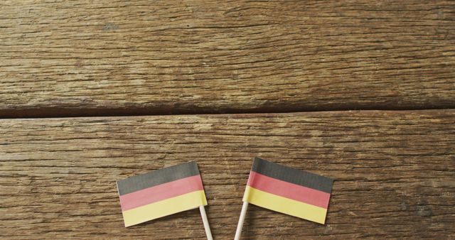 Two small German flags are lying on a wooden surface, showcasing the national colors of black, red, and gold. Ideal for use in projects related to German national holidays, cultural events, history presentations, educational materials, or patriotic celebrations. The rustic wooden texture adds a charming, vintage feel.
