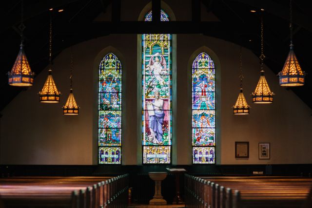 Elegant Gothic church interior featuring colorful stained glass windows, ornate chandeliers, and rows of wooden pews. Perfect for using in articles about religious architecture, historic churches, places of worship, or illustrating concepts of faith and spirituality.