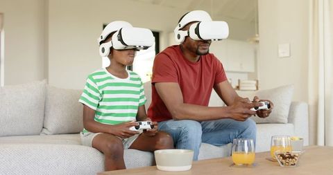 African American father and son wearing VR headsets playing video games on home sofa
