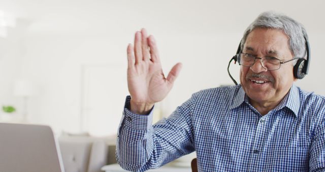 Senior man wearing headset, smiling and waving while engaging in a video call on a laptop from home. Ideal for illustrating remote communication, virtual meetings, online learning, technology usage among elderly, and work from home setups.