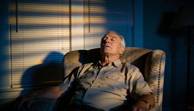 Senior man reclining in armchair at dusk with sunlit blinds casting dramatic shadows