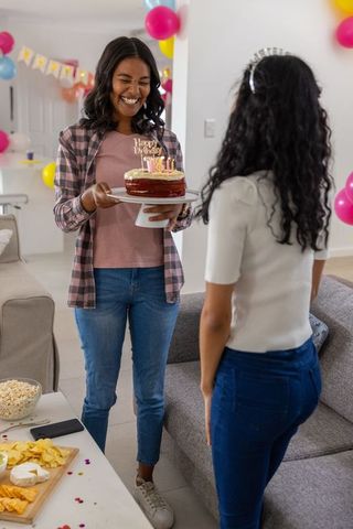 Smiling diverse mother presenting birthday cake to teenage daughter at colorful home party