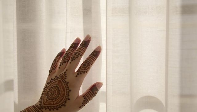 Lifting henna decorated hand revealing intricate mandala through sheer curtain backlit