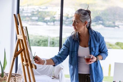 Mature woman painting at easel in bright coastal studio holding cup of tea