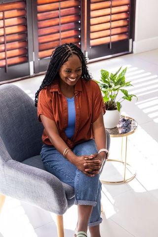 Smiling african woman relaxing in sunlit living room in gray armchair wearing casual outfit