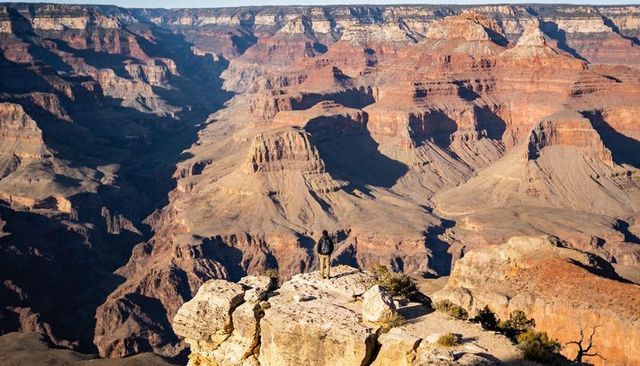 Hiker standing on cliff edge overlooking vast sunlit grand canyon layered sandstone vistas