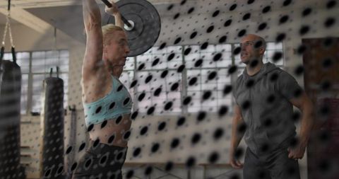 Mid-adult woman lifting loaded barbell overhead while coach observing behind mesh