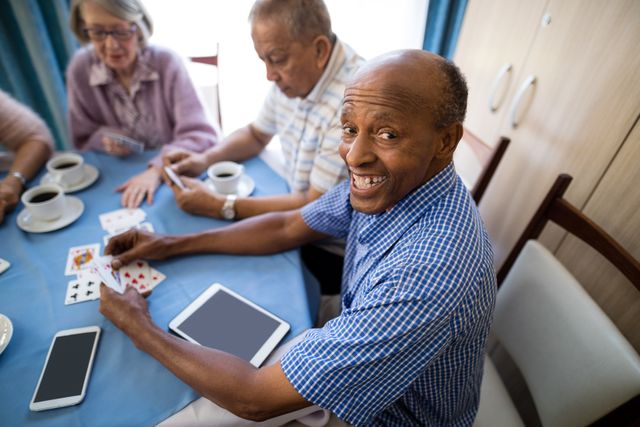 Senior man enjoying a card game with friends at a nursing home. They are sitting around a table with coffee cups and a tablet. Ideal for use in content related to elderly care, retirement communities, social activities for seniors, and promoting companionship and happiness in senior living.
