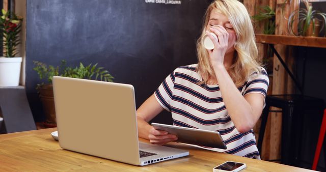Young woman with blonde hair drinking coffee while working on a tablet and using a laptop in a casual workspace. Great for illustrating concepts related to remote work, freelancing, digital nomads, modern workspaces, multitasking, and small business operations in promotional materials, website design, blog posts, and marketing activities.