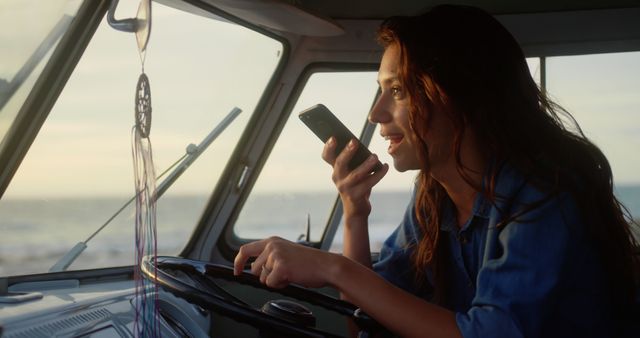 Young woman driving a classic van by the ocean, enjoying a beautiful sunset. She is engaged with her smartphone, possibly sending a voice message. Ideal for themes around travel, freedom, technology, and outdoor lifestyle advertisements or articles.