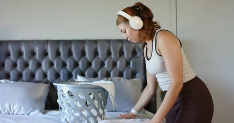 Woman folding laundry on bed while listening to headphones in minimalist neutral bedroom
