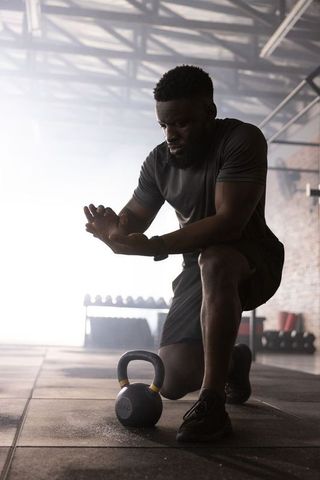 African American athlete kneeling and chalking hands with kettlebell in industrial gym