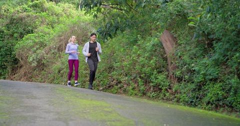 Diverse couple jogging uphill on mossy road through lush forest trail for outdoor fitness