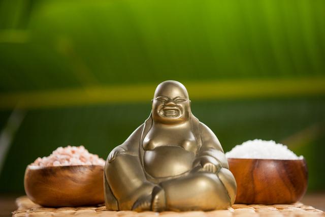 Laughing Buddha figurine placed between two wooden bowls filled with sea salt. The woven mat and green background add to the tranquil and serene setting. This photo can be used for themes related to relaxation, meditation, spirituality, wellness, and interior decor.