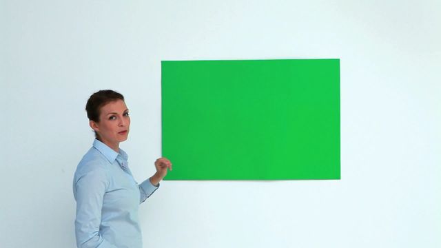 Female professional presenting green blank poster on white wall, ideal for marketing, corporate communication, or educational purposes. Useful for visual aids in business meetings, conferences, promotional events, and instructional settings.