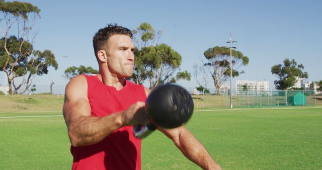 Fit caucasian man exercising outdoors, squatting and lifting kettlebell weight with one arm. cross training for fitness at a sports field.