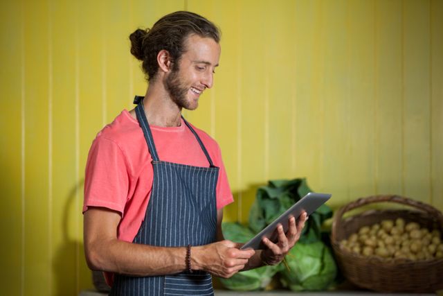 Male staff member in an organic supermarket using a digital tablet while smiling. He is wearing an apron and standing in front of fresh vegetables. Ideal for use in content related to organic food, grocery stores, customer service, small businesses, and technology in retail.