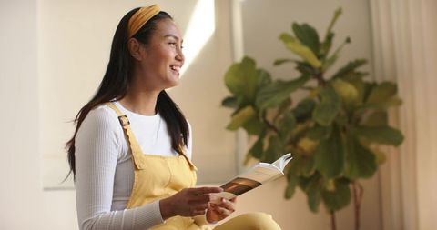 Sunlit asian woman reading and smiling in yellow overalls and headband, minimalist home interior