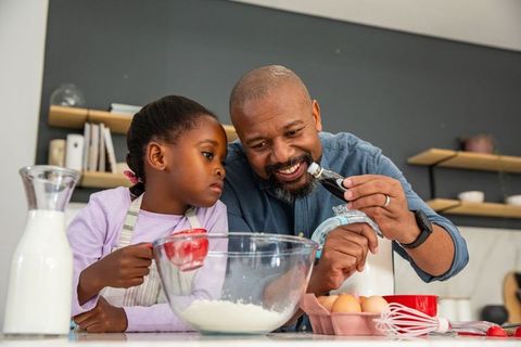 African american father baking with daughter measuring flour and adding vanilla in kitchen