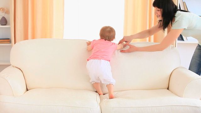 A mother is gently supporting her baby as they crawl on a white couch, showcasing a caring bonding moment. This scene highlights parenting and child development in a warm family setting, suitable for themes of nurturing, early growth, and home life.