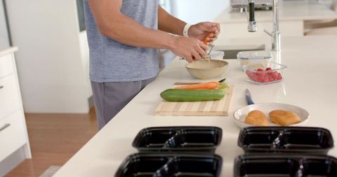 Healthy meal prep routine: man peeling carrot and arranging cucumber, containers on countertop