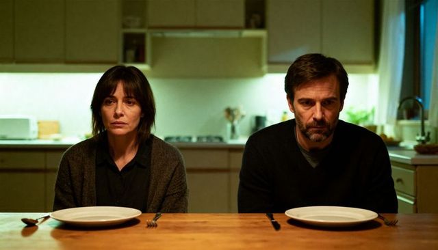 Midlife couple sitting apart at kitchen table with empty plates showing domestic tension, moody