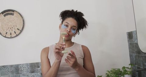African american woman using jade facial roller and under-eye patches for skincare routine