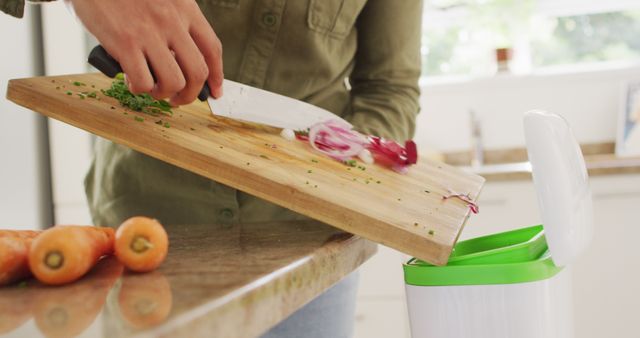 This image shows a person transferring food waste from a cutting board into a compost bin in a kitchen. Suitable for content regarding sustainable living, composting practices, and environmental cleanliness. Perfect for articles, blogs, and guides on reducing household waste, zero-waste lifestyles, and eco-friendly habits.