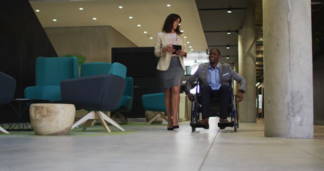 Businesswoman in professional attire walking alongside a male colleague in a wheelchair in the modern office lobby, representing diversity and inclusion in the workplace. Suitable for themes such as office collaboration, teamwork, accessible work environments, and corporate employee relations.