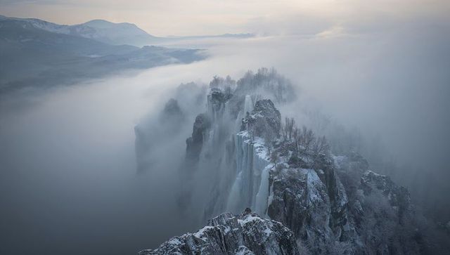 Frozen mountain ridge emerging from fog with icy waterfalls and snow-covered cliffs at dawn