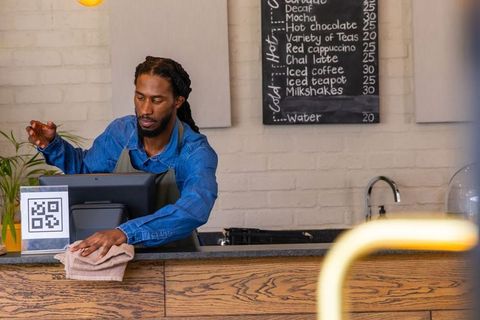 African american barista wiping counter while using touchscreen pos and qr code at cafe