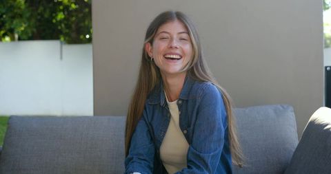 Young woman laughing on backyard patio sofa wearing denim shirt and cream top