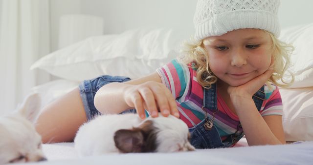 Little girl is gently petting two small, cute puppies while laying on a bed in a cozy bedroom. She is wearing a white knit hat and striped shirt. Ideal image for themes like childhood, pet care, family, and coziness.