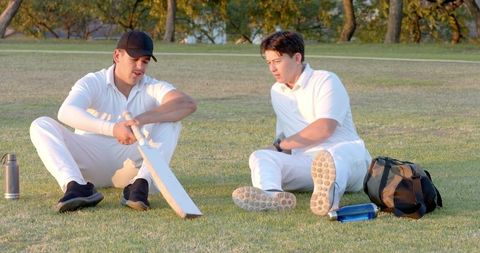 Cricket teammates sitting on grass examining bat at golden hour, relaxing after match