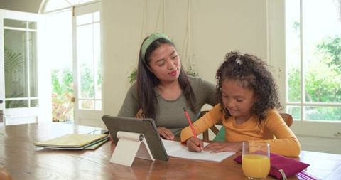 Mother helping daughter with homework using tablet and pencil at sunny home table