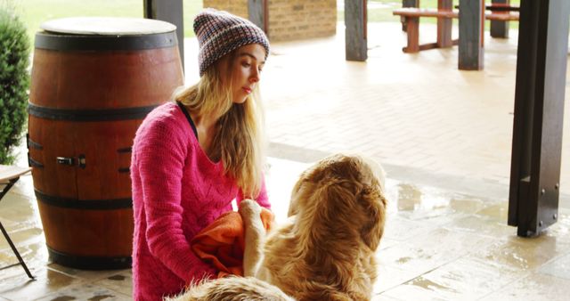 Woman wearing pink sweater and beanie, attending to her golden retriever with a towel on covered porch during rainy weather. Suitable for themes like pet care, rainy days, outdoor activities with pets, and lifestyle photography. Ideal for websites, blogs, and advertisements focused on pet care, animal companionship, and rainy day activities.