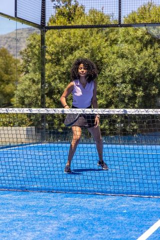 African american woman playing padel at net on bright blue court wearing lavender top