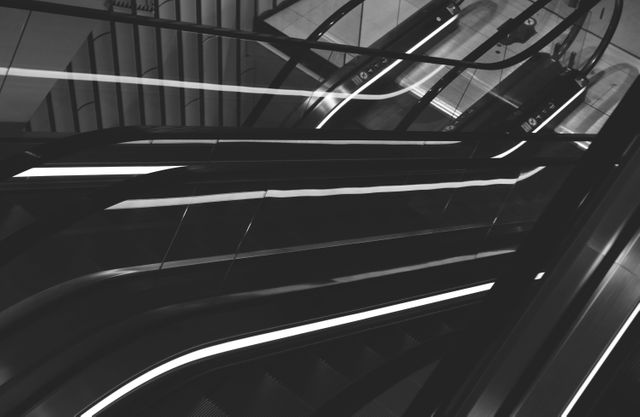 Black and white image showing a modern escalator in a shopping mall setting. The sleek lines and reflective surfaces create a minimalist and urban feel. Ideal for use in articles about urban living, transportation, architecture, and modern public spaces. Suitable for enriching content related to advanced design and infrastructure images.