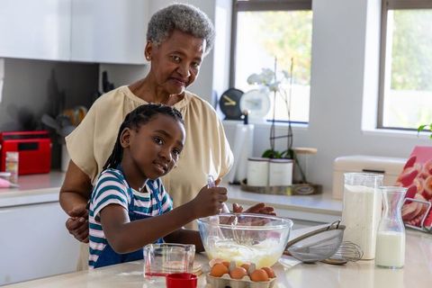 Grandmother guiding granddaughter whisking batter in bright modern kitchen during baking lesson
